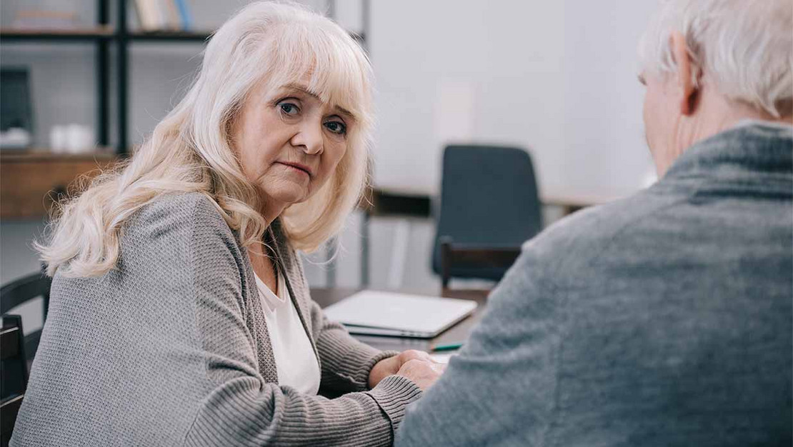 Eine ältere Frau sitzt mit ihrem Mann am Tisch und blickt besorgt in die Kamera. 